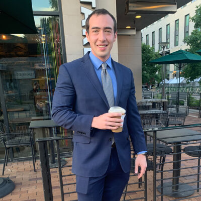 Kai Bidell has short dark hair and wears a navy blue suit with a blue dress shirt and gray tie. Bidell stands in front of a Starbucks while holding an iced coffee.