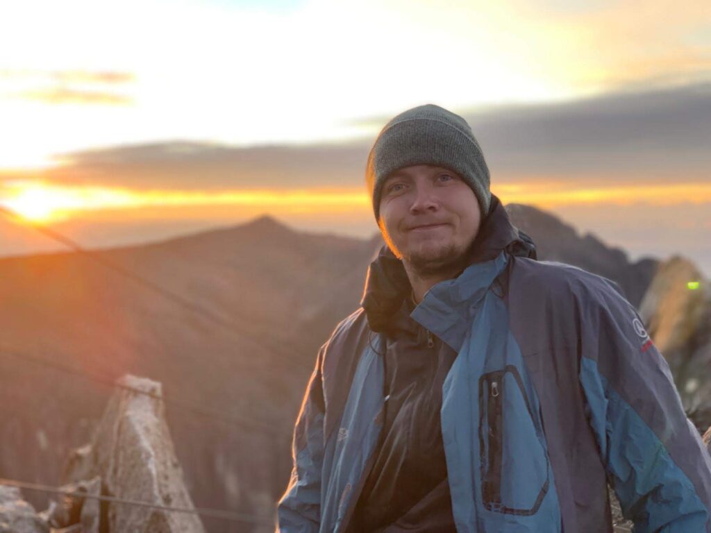 Peter Hansen stands on Mt. Kinabalu with a golden orange sunset in a cloudy sky and mountains behind him. He wears a gray beanie and a dark and light blue windbreaker jacket.