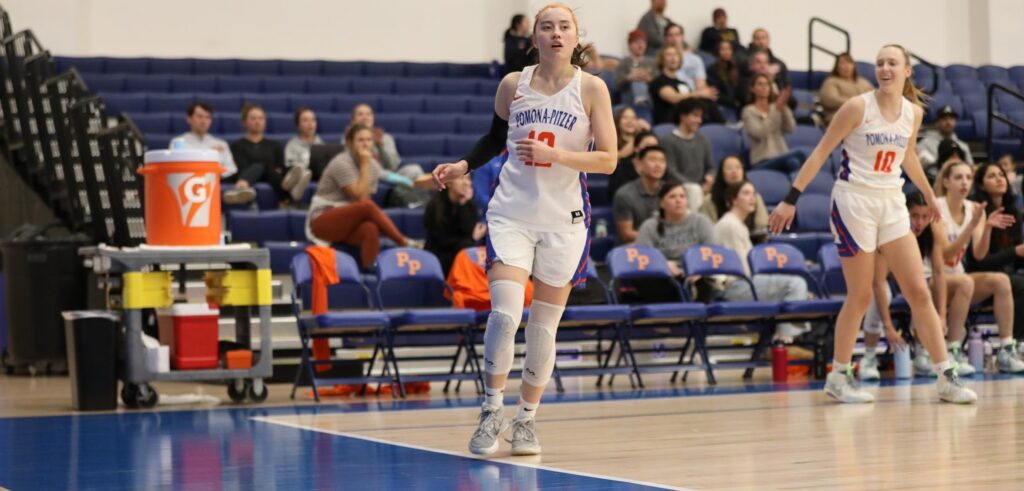 Madison Quan wears her long dark hair in a ponytail and a white basketball uniform with Pomona-Pitzer in blue text and 12 in orange text on the front. She jobs across the court against a backdrop of a blurred out crowd in the navy blue bleachers.