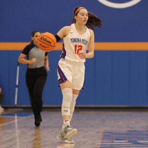 Madison Quan wears her long dark hair in a ponytail and a white basketball uniform with Pomona-Pitzer in blue text and 12 in orange text on the front. She dribbles the basketball on the court as a player in a black and yellow uniform tries to block her.