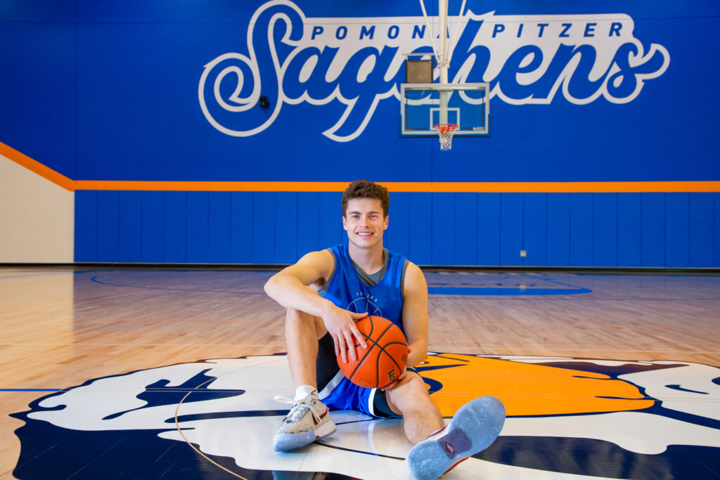 Peyton Mullarkey holds a basketball between his hands as he sits on the Cecil the Sagehen logo in the middle of the basketball court. He has short brown hair and wears a blue practice uniform and gray sneakers. Behind him is a basketball net and a blue wall with Pomona-Pitzer Sagehens in blue text with a white outline. Pomona-Pitzer is in smaller plain script and Sagehens is in large cursive script.