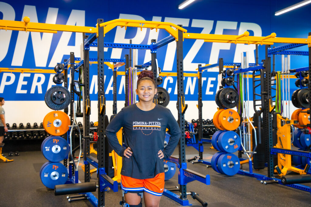 Charlize Andaya stands with her hands on her hips in front of the orange and blue Olympic weights in the Athletic Performance Center. She has long dark brown hair with reddish tips pulled into a bun on top of her head and wears a long-sleeved black shirt with Pomona-Pitzer Basketball in white text and orange shorts with a white stripe in between two blue stripes at the bottom.