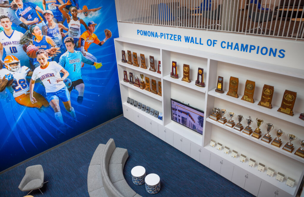 An angled photo of the first floor taken from the second floor. On the left is a photo collage of Sagehens student-athletes playing basketball, football, soccer, baseball, swimming, and other sports against a blue backdrop. On the right is white shelf full of trophies with blue text above it that reads Pomona-Pitzer Wall of Champions. A comfortable gray couch and chair are scattered across dark blue striped textured carpet.
