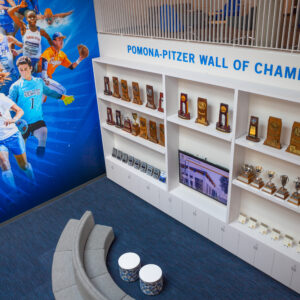 An angled photo of the first floor taken from the second floor. On the left is a photo collage of Sagehens student-athletes playing basketball, football, soccer, baseball, swimming, and other sports against a blue backdrop. On the right is white shelf full of trophies with blue text above it that reads Pomona-Pitzer Wall of Champions. A comfortable gray couch and chair are scattered across dark blue striped textured carpet.