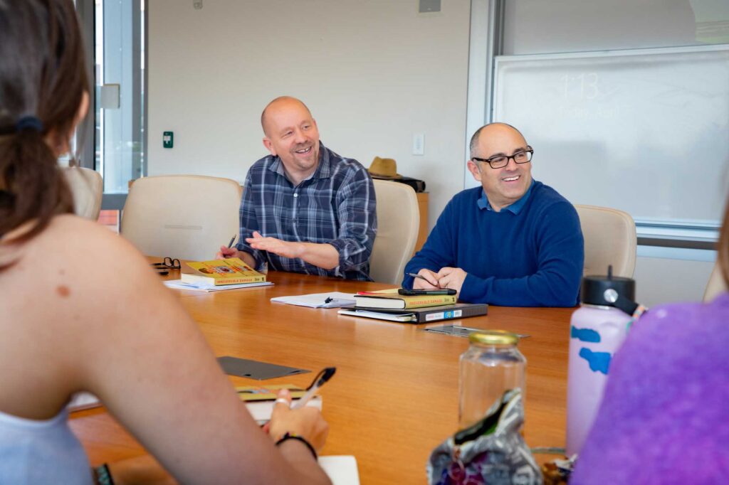 Jon Shields and Phil Zuckerman sit beside each other at a long wood table. They smile and look to their left at a student offscreen. Shields is bald and wears a long-sleeved dark blue and white plaid shirt. Zuckerman is bald and wears black glasses and a blue sweater. Copies of the book Irreversible Damage are in front of each of them.