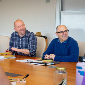 Jon Shields and Phil Zuckerman sit beside each other at a long wood table. They smile and look to their left at a student offscreen. Shields is bald and wears a long-sleeved dark blue and white plaid shirt. Zuckerman is bald and wears black glasses and a blue sweater. Copies of the book Irreversible Damage are in front of each of them.