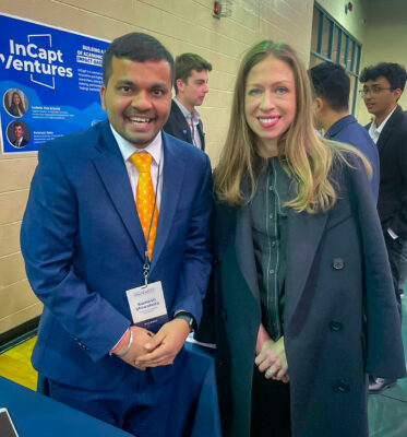 Sumesh Shiwakoty stands beside Chelsea Clinton while a crowd is mingling behind. Shiwakoty wears a dark blue blazer, white collared shirt, and orange tie and has short dark hair. Clinton has long blond hair and wears a black button shirt and black coat over her shoulders.