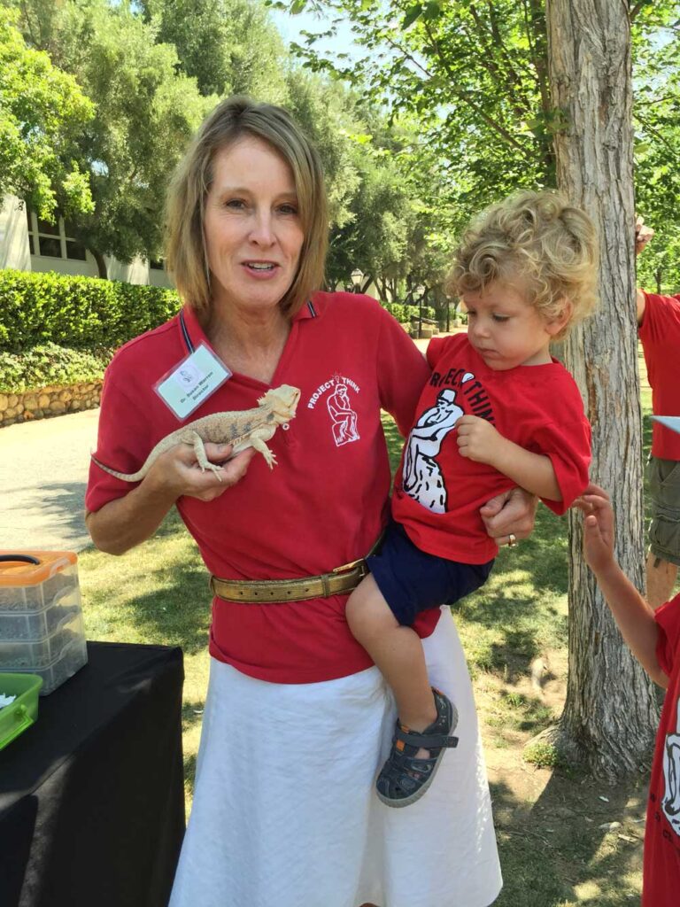 Susan Warren holds her toddler grandson on her hip with one arm and holds a pale yellow reptile in her other hand. Warren and her grandson wear red Project think shirts with the logo of a line drawing of The Thinker sculpture. Warren has shoulder-length blond hair and her grandson has curly blond hair.