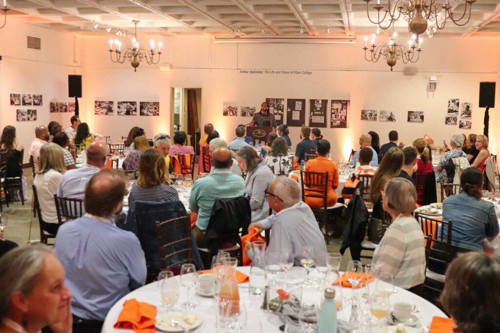 Sekou Andrews stands at a Pitzer College podium while speaking to a room full of alumni. They are gathered around tables covered in white tablecloth, orange napkins, and glasses in a warmly lit room with chandeliers hanging above.