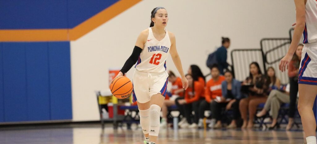 Madison Quan '22 bounces a basketball on the court and wears a white uniform with Pomona-Pitzer in blue text and the number 12 in orange text. Quan has long dark brown hair in a ponytail.
