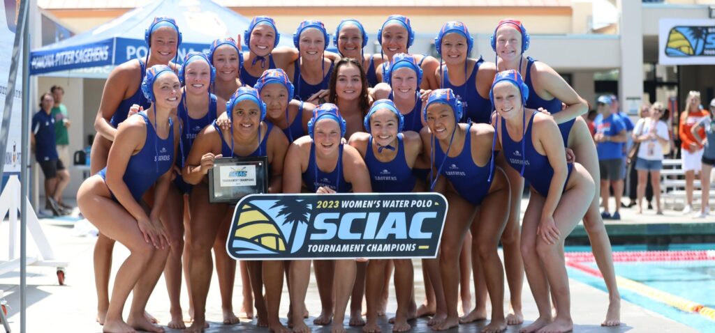 The women's water polo team poses by the poolside with a sign that reads 2023 Women's Water Polo SCIAC Tournament Championships. They wear blue swim caps and bathing suits.