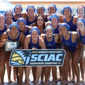 The women's water polo team poses by the poolside with a sign that reads 2023 Women's Water Polo SCIAC Tournament Championships. They wear blue swim caps and bathing suits.