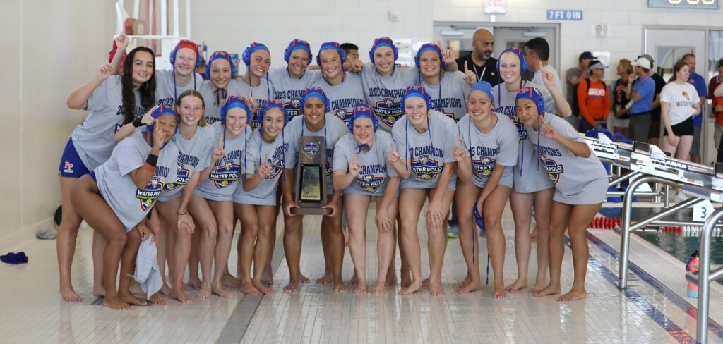 The women's water polo team pose by the poolside with the trophy in the middle. They wear blue swim caps and gray T-shirts with the championship logo over their blue swimsuits. All of them raise their forefinger in a number one sign.