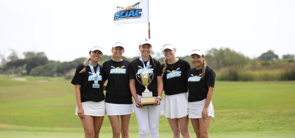 The women's golf team stands in a green golf course with the white SCIAC flag flying behind them and one member holding a trophy in the middle. They wear black T-shirts with the light blue SCIAC logo and white skirts and pants.