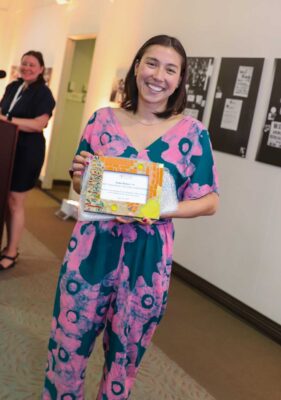 Keiko Budech wears a teal and pink jumpsuit as she stands and holds a colorful frame with her alumni award certificate. Budech has straight shoulder-length brown hair.