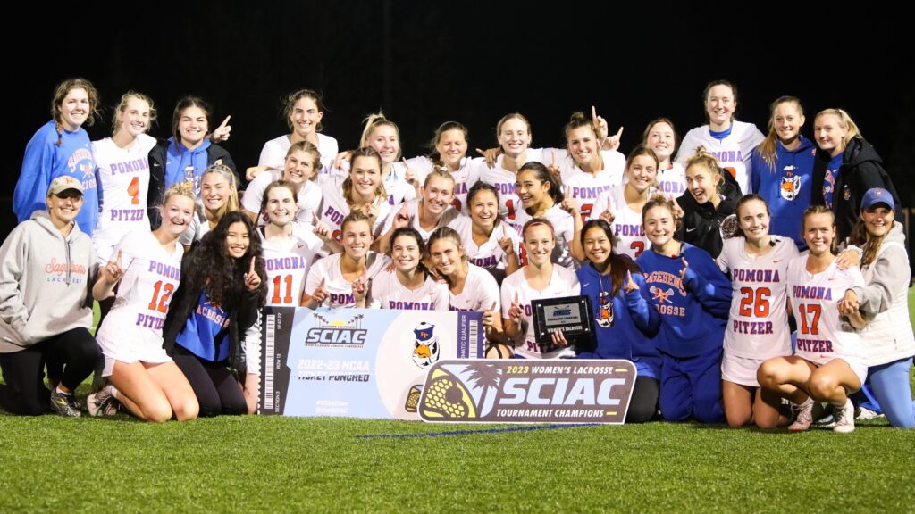 The women's lacrosse team gather on a green field with a sign in the middle that reads 2023 Women's Lacrosse SCIAC Tournament Champions in the middle. The team wears white uniforms with blue and orange lettering and blue uniforms with orange lettering.