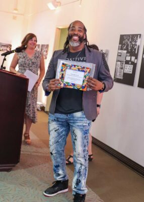 Sekou Andrews wears a gray jacket over a black T-shirt and faded jeans with various words and patterns written in blue and black ink. He stands and holds a colorful frame with his alumni award certificate. Andrews has shoulder-length dark brown hair and a beard with dark brown and gray hair.