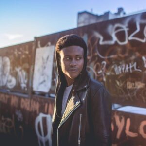 Tyriek White wears a black leather jacket with a black hood over his head while he stands in front of a rust brown wall with graffiti writing.