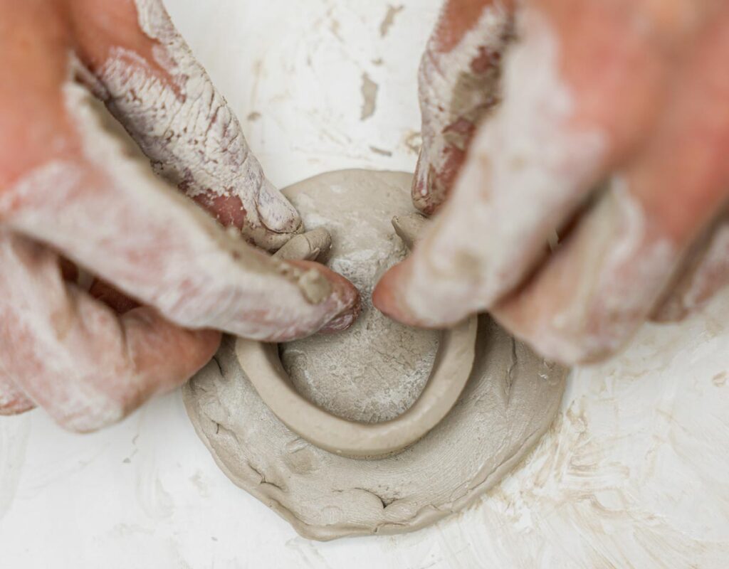 Two hands coated with clay take two ends of a thin roll of clay in the shape of a circle to connect the ends.