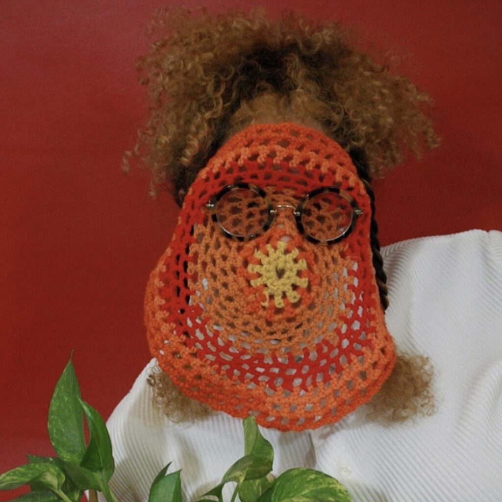 Jaspa Ureña stands behind a leafy green plant and has an orange, red, and yellow crocheted circle and a pair of round black glasses over their face. Ureña has curly light brown hair and wears a white sweater. 

