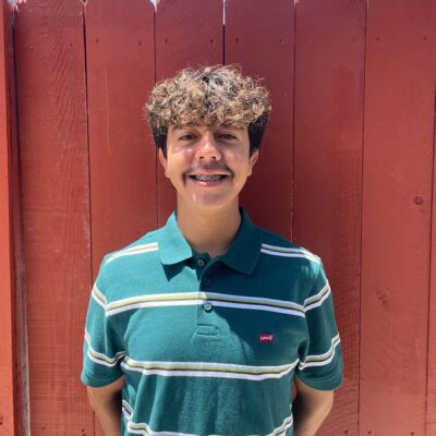 Jesus Ceja has short curly brown hair and wears a green collared shirt with horizontal white stripes. Ceja stands in front of a reddish brown fence.