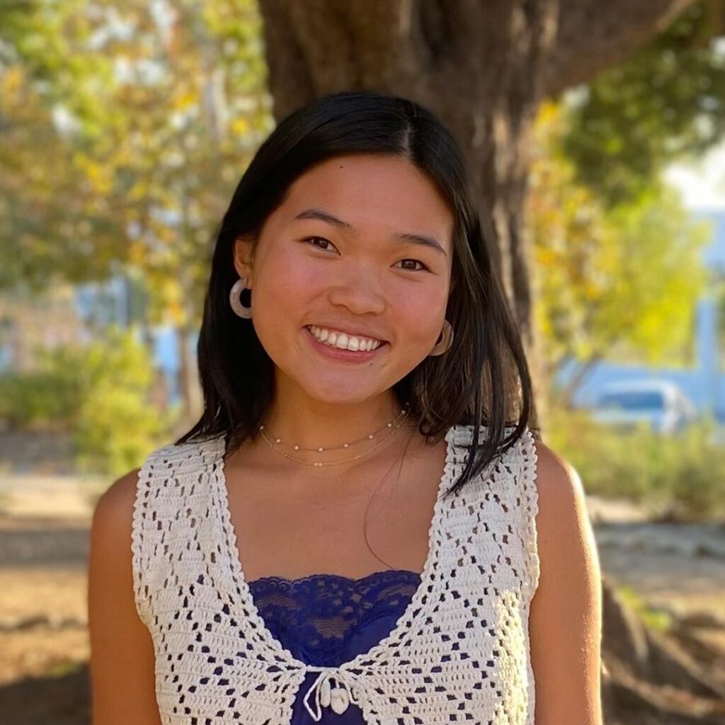 Keely Nguyen has long straight black hair and wears hoop earrings and a knitted white top over a blue top while standing in front of a tree that is out of focus.