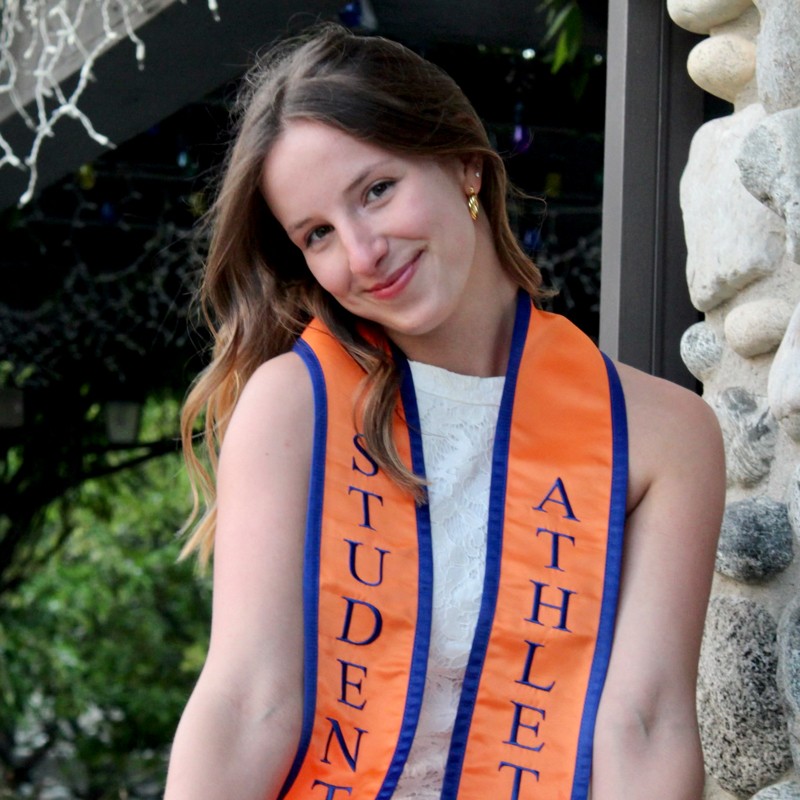 Morgane Monjauze has long wavy brown hair with blond tips and wears an orange and blue student athlete graduation stole over a white lace dress.