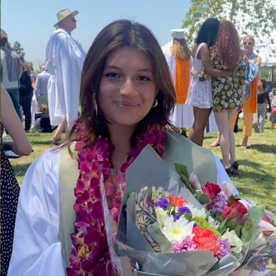 Olivia Rosenberg-Chávez has shoulder-length straight brown hair and wears white graduation regalia and a purple lei while holding a flower bouquet.