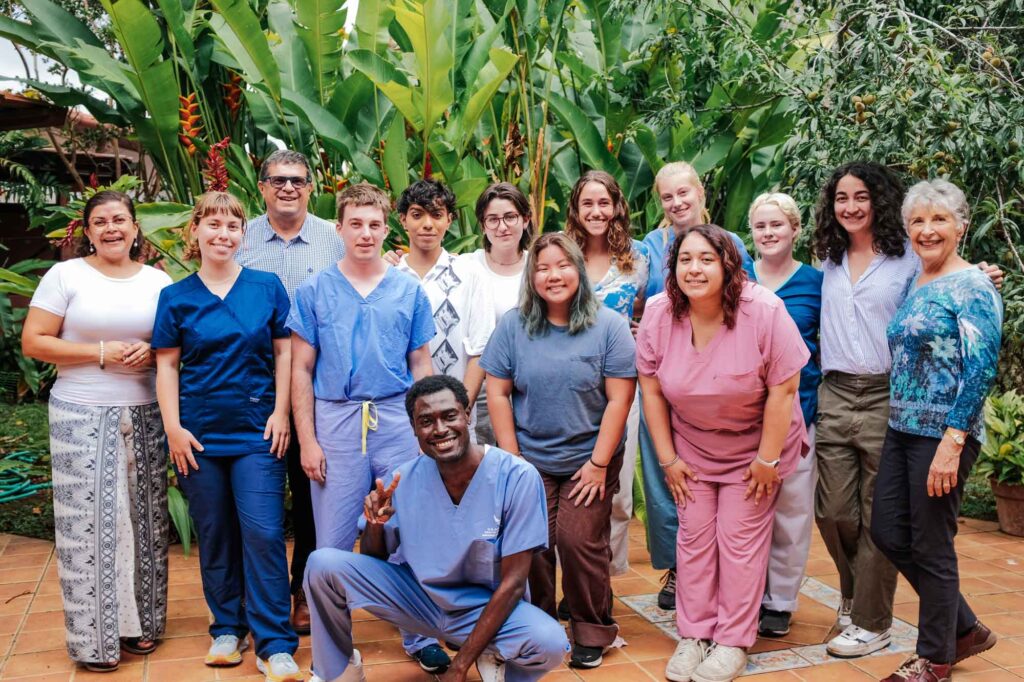 Pitzer students pose in a group photo with Ann Stromberg on the far left and with the ICADS staff with green tropical plant life behind them.