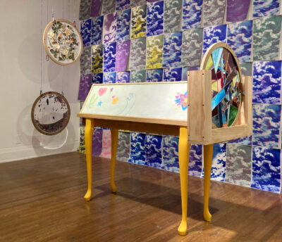 A giant kaleidoscope stands on four yellow legs and has a disc at the end made of collaged glass fragments in different colors. Two more discs with glitter and with plant materials hang from the ceiling. Behind the kaleidoscope is a wall of blue, gray, and yellow prints of a photo of a cloud.