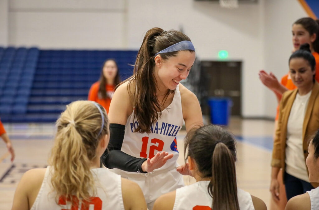 Madison Quan has long straight dark hair in a ponytail and wears a blue headband and a white basketball uniform with the blue text that says Pomona-Pitzer and orange text that says 12. Quan smiles while extending hands for a high five to two of her teammates sitting on a bench.