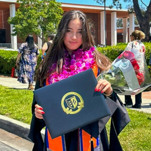 Madison Quan wears black graduation robe with a purple lei, a gray stole, and an orange and blue stole. Quan holds up the diploma from CGU and tucks a bouquet of red roses in her left arm. Quan has long straight dark brown hair.