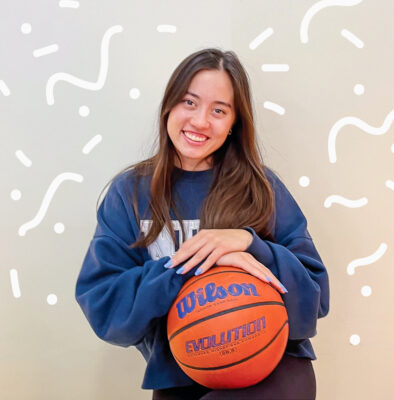 Madison Quan has long straight dark brown hair and wears a dark blue sweatshirt. Quan sits with her arms crossed over a basketball. White squiggle marks surround her.