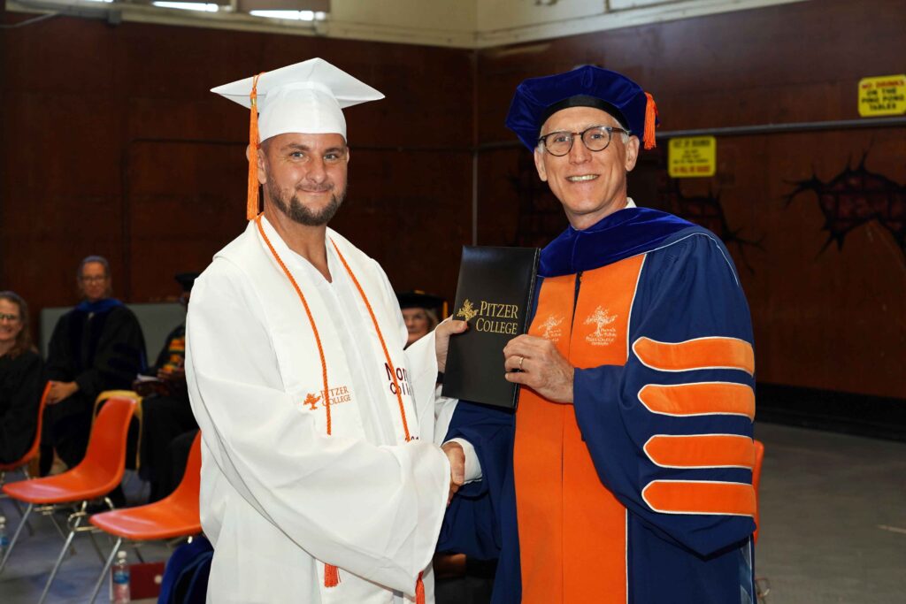 Jennings wears white and orange regalia and Thacker wears dark blue and orange regalia. Jennings shakes Thacker's hand as Thacker hands over a black Pitzer College diploma.