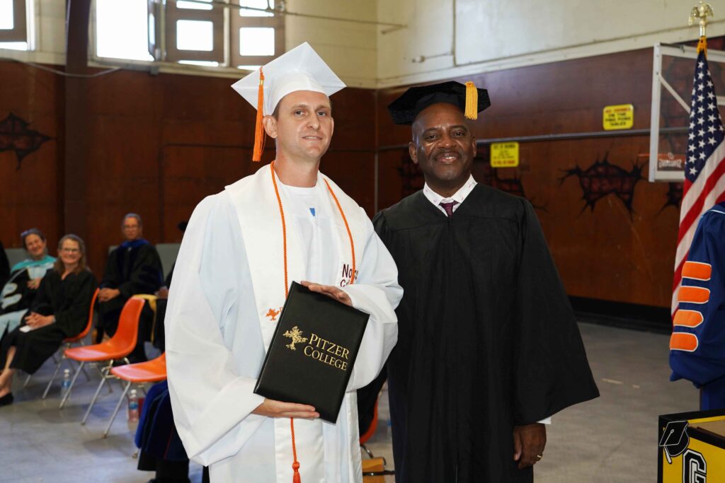 Rose wears white and orange regalia and Foster wears black regalia as they stand side by side. Rose holds up a black Pitzer College diploma.