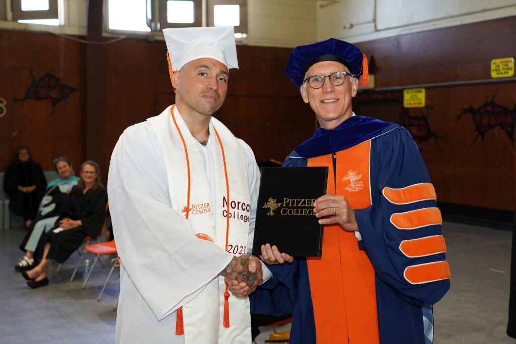 Rivera wears white and orange regalia and Thacker wears dark blue and orange regalia. Rivera shakes Thacker's hand as Thacker hands over a black Pitzer College diploma.