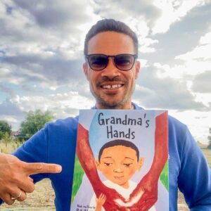Joel Harper holds up the book Grandma’s Hands with one hand and points to the cover with the other hand. The front cover features an illustration of a little boy with his grandmother’s arms embracing him. Harper has short dark hair and wears sunglasses and a long-sleeved blue shirt.