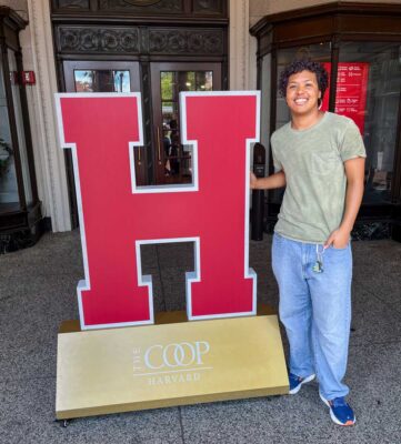 Cooper stands next to a giant red H sign on top of a gold stand with white text that reads The COOP Harvard. Cooper wears a light green T-Shirt, light blue pants, and dark blue sneakers.