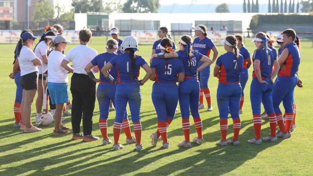 The women's softball team stand in a circle with their coaches on a field. The team wears blue Sagehens uniforms with orange and white socks.