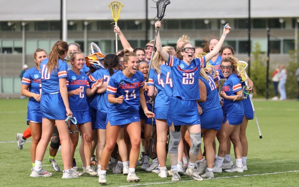 The women's lacrosse team embrace each other and cheer victoriously on the field. They wear blue Sagehens uniforms with white and orange accent colors.