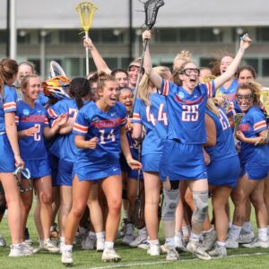 The women's lacrosse team embrace each other and cheer victoriously on the field. They wear blue Sagehens uniforms with white and orange accent colors.