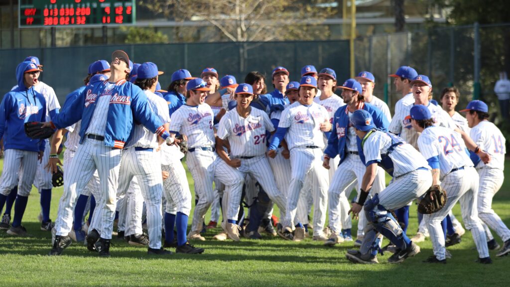 The men's baseball team gather on a field to cheer in victory. They wear white uniforms with blue and orange hats and long-sleeved blue shirts