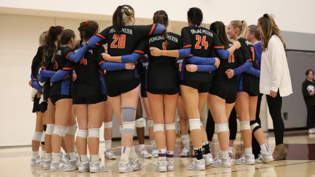The women's volleyball team stand in a circle on a court. They wear black uniforms with orange, white, and blue accent colors and white knee pads.