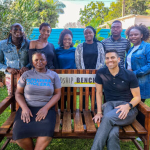 Marcus Rodriguez on the far right sits on a wood bench with a sign for Friendship Bench on the back rest. A Friendship Bench worker sits on the opposite side of the bench and other workers gather behind the bench.