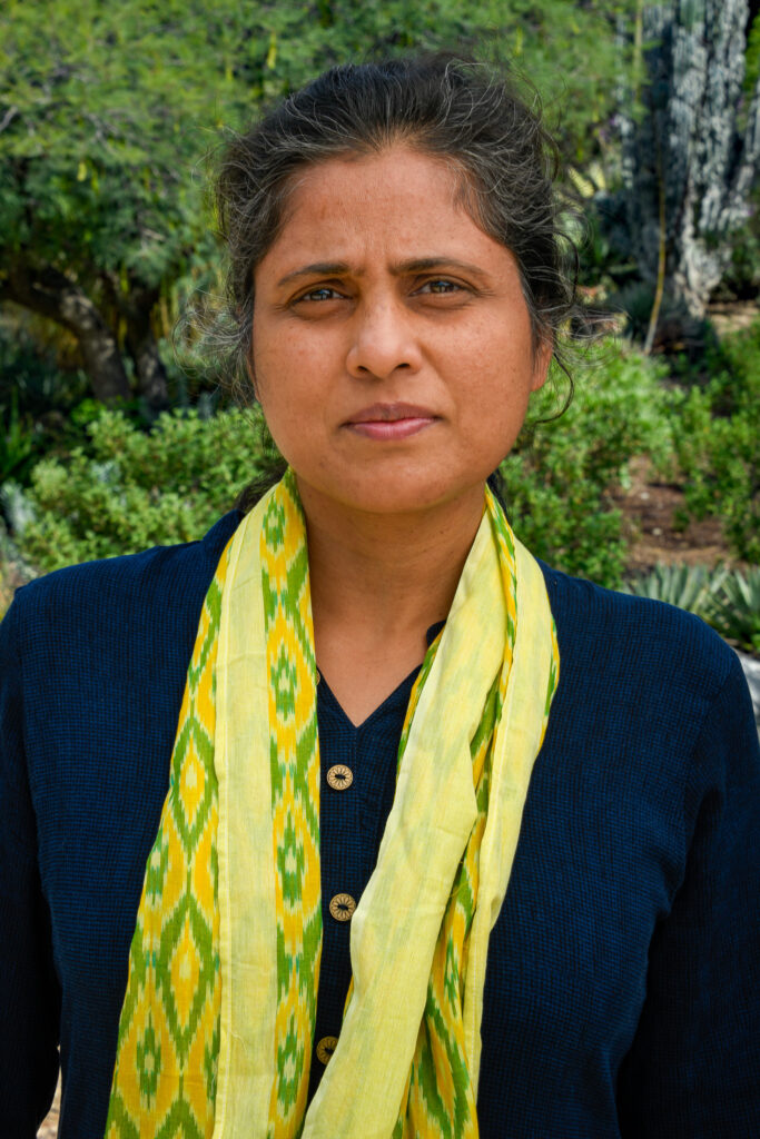 Deepti Goes has dark hair pulled back and wears a dark blue long-sleeved shirt and a yellow and green scarf with diamond patterns.