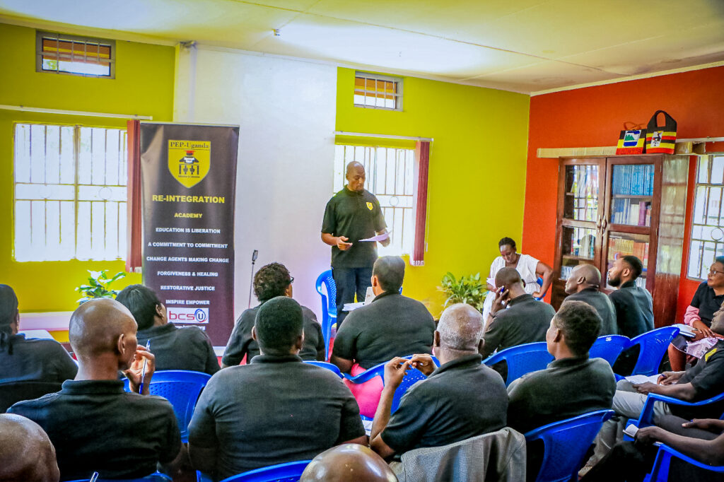 Kenneth Butler stands in front of a class while reading from a paper in his hand. Butler wears a black polo shirt with the yellow logo for PEP-Uganda on the upper left corner. To his right is a black banner for the Reintegration Academy with the statement “Education is liberation, a commitment to commitment, change agents making change, forgiveness & healing, restorative justice, inspire empower.” 