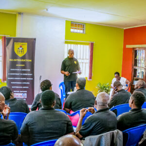 Kenneth Butler stands in front of a class while reading from a paper in his hand. Butler wears a black polo shirt with the yellow logo for PEP-Uganda on the upper left corner. To his right is a black banner for the Reintegration Academy with the statement “Education is liberation, a commitment to commitment, change agents making change, forgiveness & healing, restorative justice, inspire empower.”