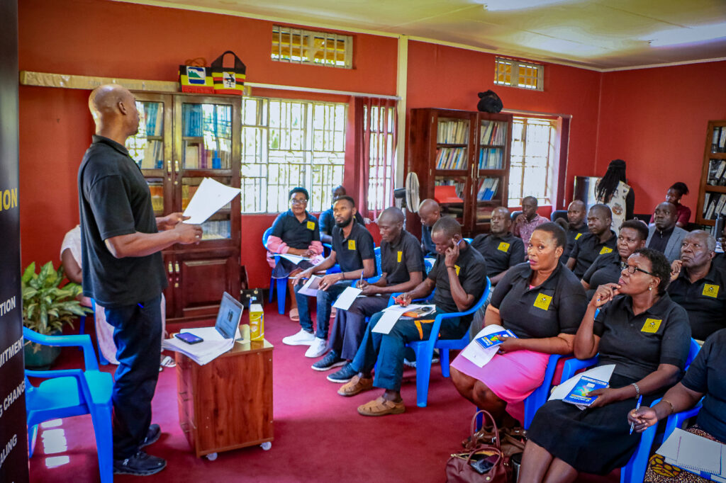 A classroom full of people in black polo shirts with the yellow logo for PEP-Uganda sit at attention as Butler presents to them.