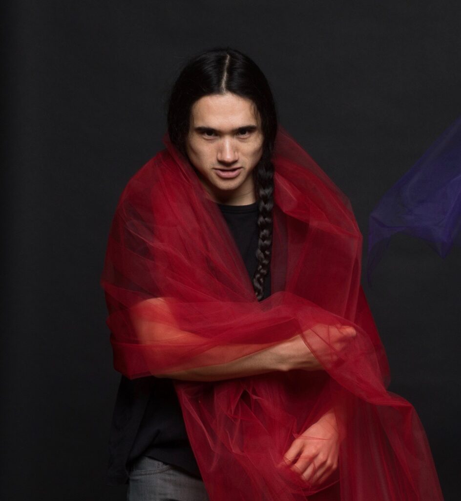Micah Huang has long black hair in a braid and wraps red tulle fabric around his shoulders while looking intensely at the camera. Huang is wearing a black shirt and jeans and stands against a dark gray background.