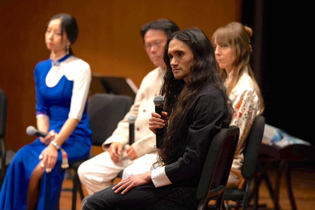 In the foreground Micah Huang is sitting in a chair and holding a mic, while in the background three other people who are out of focus also sit on chairs. Huang has long dark wavy hair and wears a long black shirt with white cuffs.
